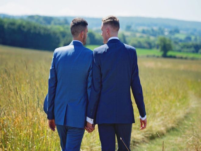 Casal homoafetivo: 2 homens de terno de mãos dadas andando pelo campo