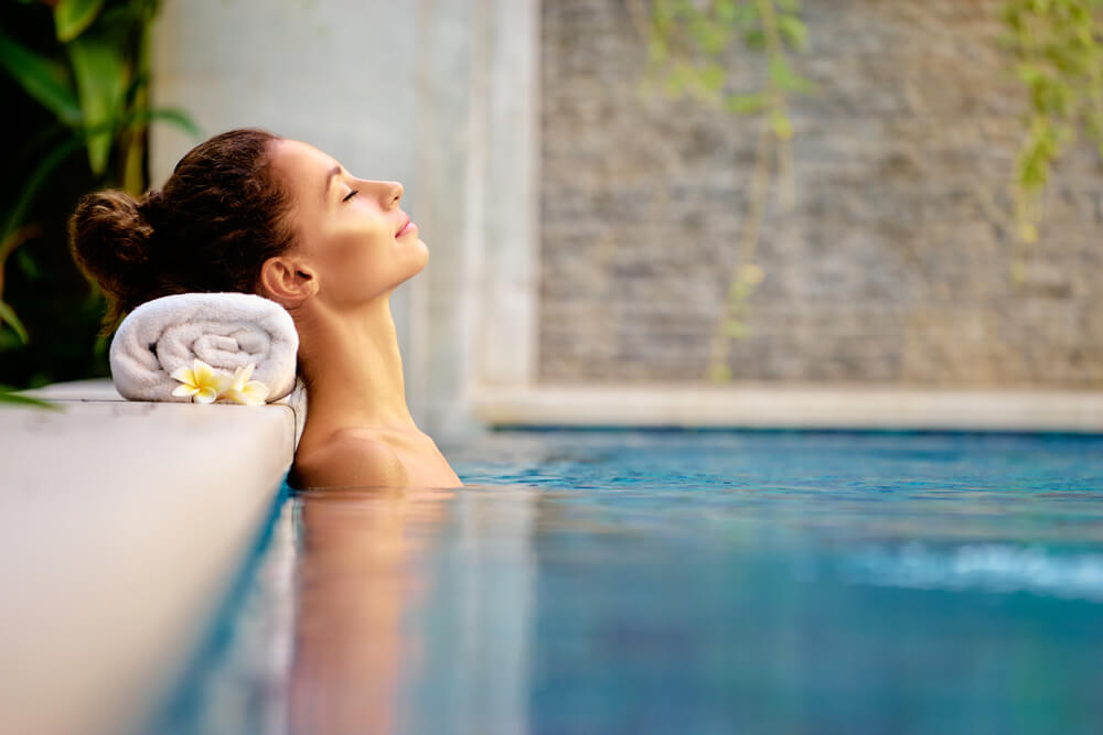 Mulher relaxa numa piscina com cabeça apoiada numa toalha branca enrolada na borda.