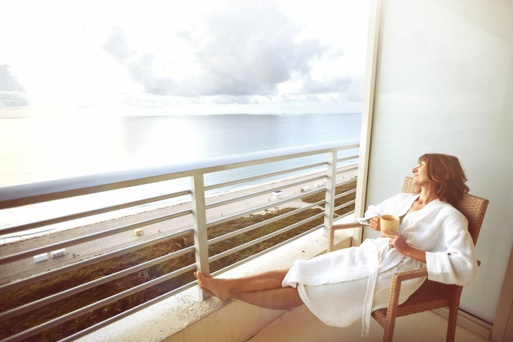 Mulher de roupão relaxa em cadeira na sacada de quarto de hotel com vista pro mar