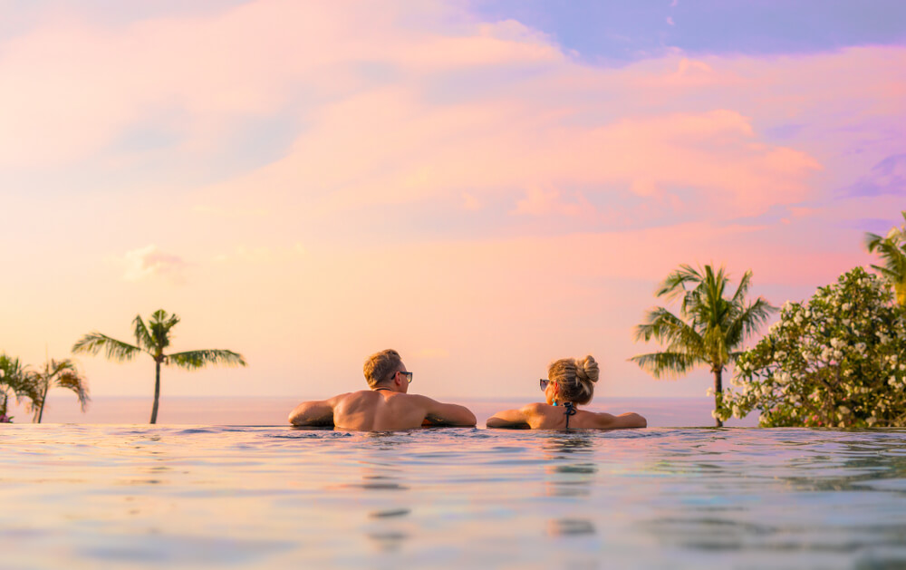Casal relaxa em borda de piscina infinita ao por do sol.