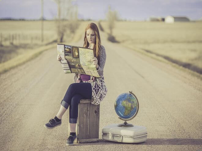 mulher na estrada sentada na mala lendo sobre viagem com  globo ao lado