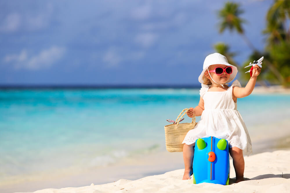 Criancinha sentada em cima de mala infantil na areia de praia brincando com um avião.
