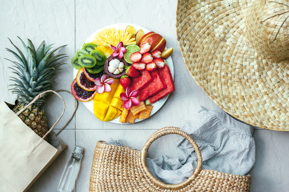 Prato com várias frutas tropicais no centro da foto rodeado de um chapéu e bolsa de palha e uma sacola de papelão com um abacaxi dentro.