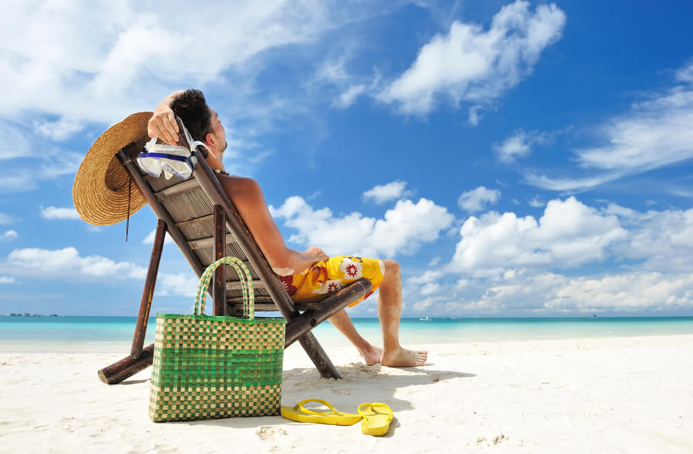 Homem de bermuda amarela relaxa em espreguiçadeira na areia de praia.