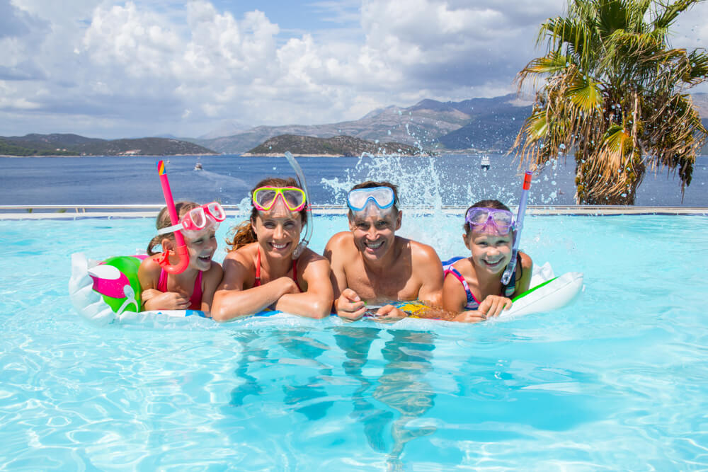 2 adultos e 2 crianças posam com boias na piscina com mar ao fundo.
