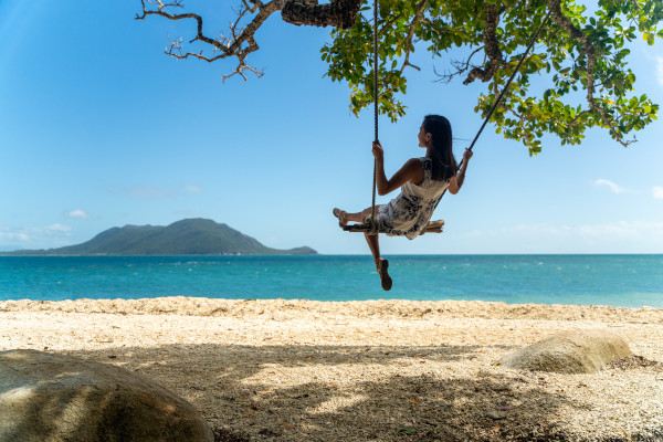 Person swinging from a tree near pristine oceans at an Australian holiday destination. The pristine waters and warm beaches of north Queensland are what make it such an attractive holiday destination. Image credit: Tourism and Events Queensland